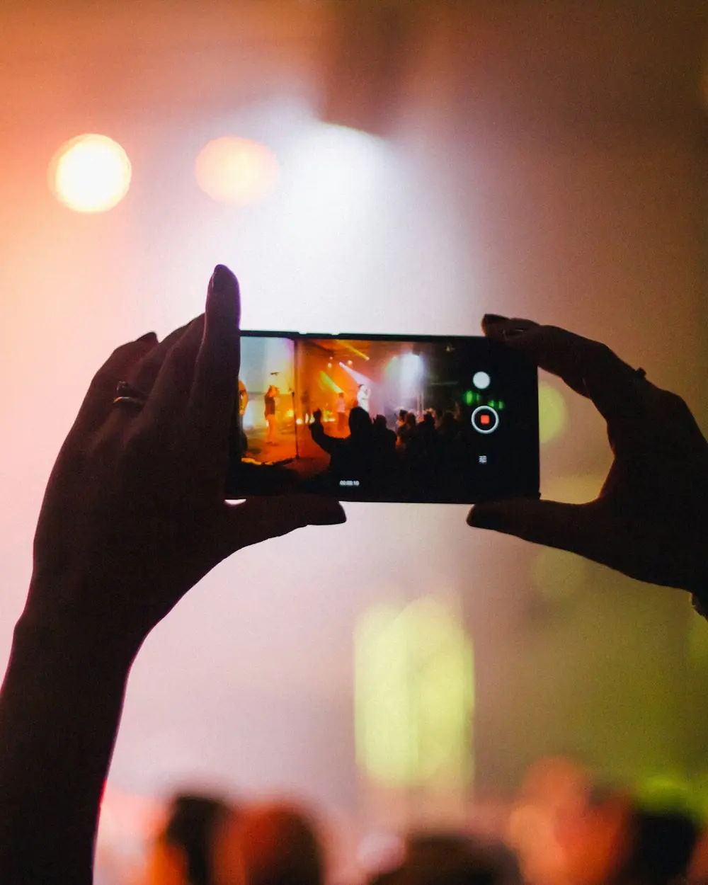 A fan holding up a phone to film a live concert, the crowd silhouetted against stage lights