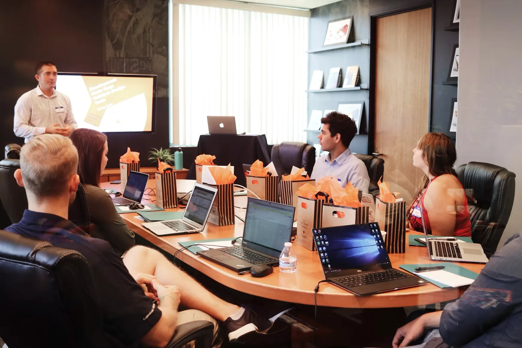 A presenter leading a session in a boardroom while colleagues follow along on laptops around the table