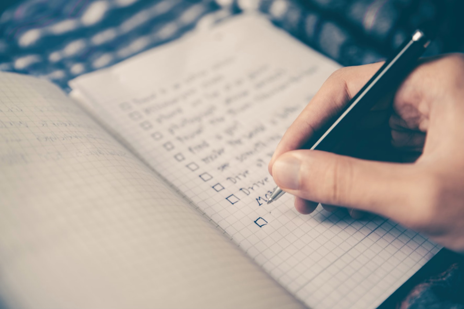 A hand writing a to-do checklist with empty tick boxes in a grid-paper notebook