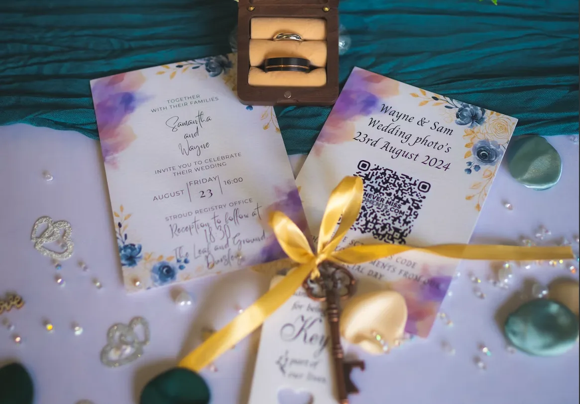 A floral wedding invitation beside a matching card with a QR code guests scan to share their photos, next to the wedding rings